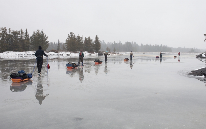 a line of cross country skiers, each pulling small sleds, make their way across a frozen lake